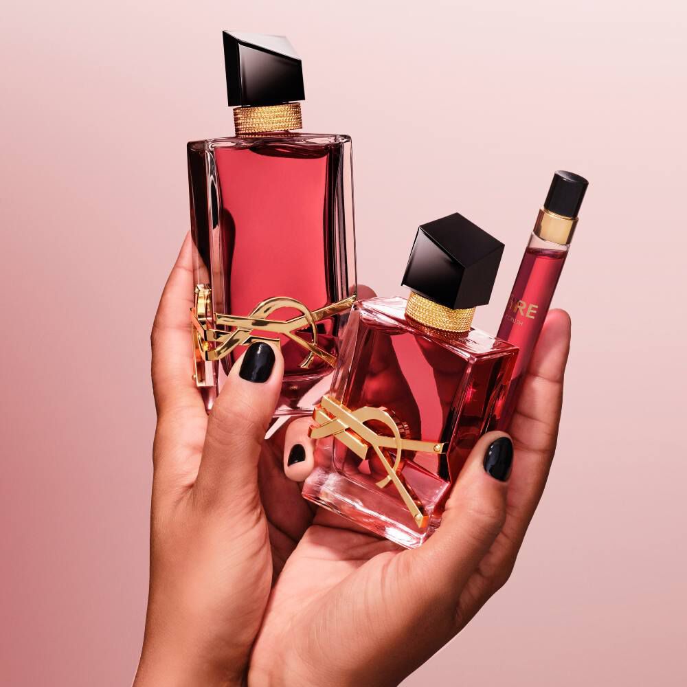 Hande mit schwarzem Nagellack halten drei verschiedene Großen von YSL Libre Parfumflaschen vor einem rosa Hintergrund.