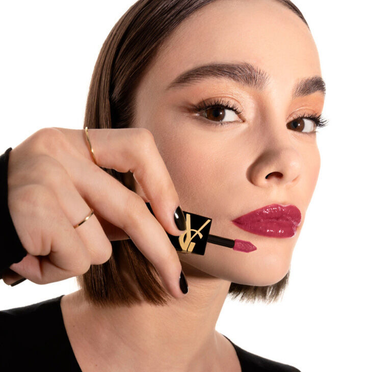 A close-up of a woman applying deep berry lipstick using a lip applicator, with a focused expression and stylish makeup. She has short, straight hair and is wearing a black top.