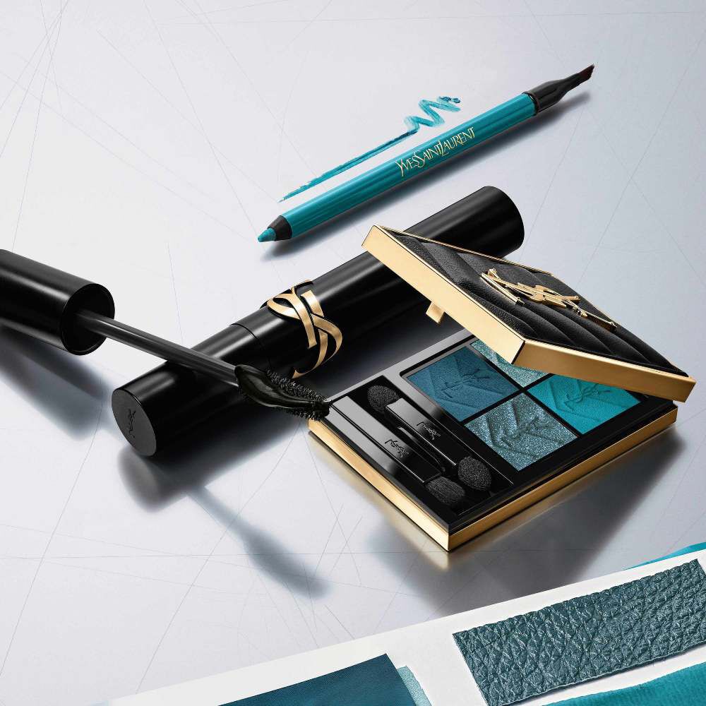 A luxury YSL beauty flat lay featuring an open gold-trimmed eyeshadow palette with blue and teal shades, a black mascara wand with product, and a teal eyeliner pencil, all arranged on a reflective silver surface.​