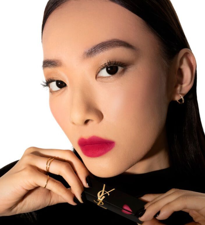 A close-up of a woman with dark hair and bold makeup, holding a Yves Saint Laurent lipstick in her hands. She has bright pink lips and a confident expression, set against a white background.