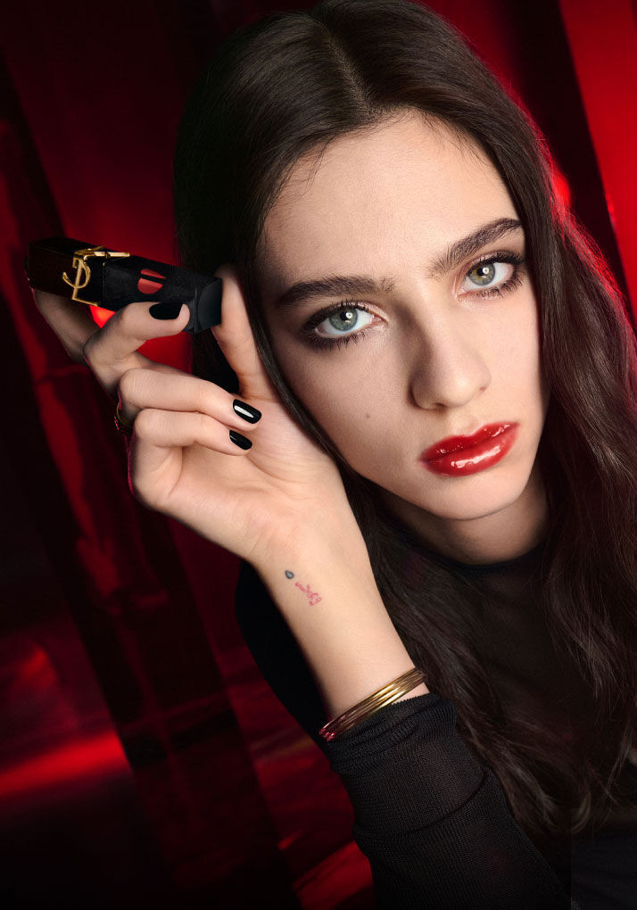 A woman with long dark hair and striking makeup holds a lipstick in her hand, staring directly at the camera against a vibrant red background. Her nails are painted black, and she wears a fitted black top, exuding a bold and confident vibe.