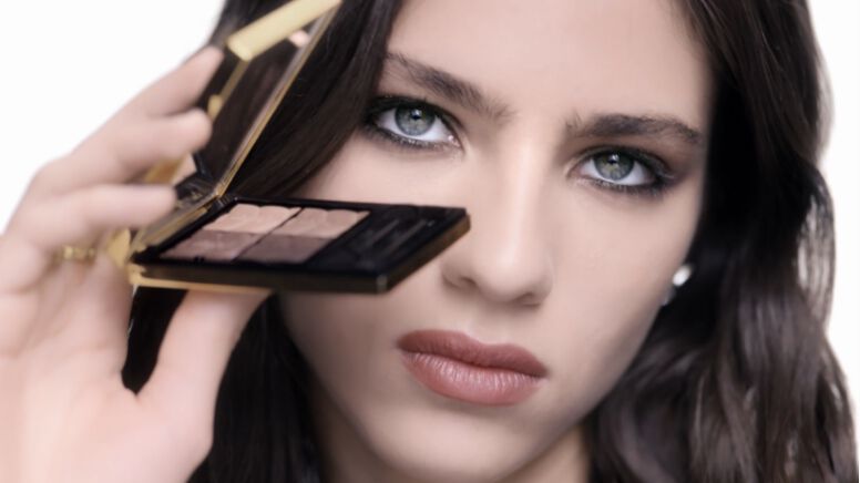 A close-up of a young woman holding a compact with eyeshadow while looking directly at the camera, showcasing her striking features and makeup.