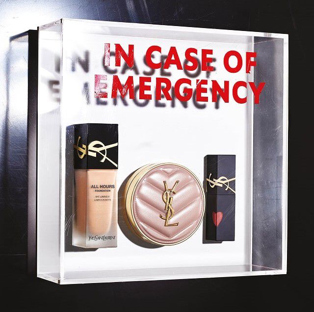 Eine transparente Plexiglasbox enthalt drei YSL Beauty Make-up-Produkte, ahnlich einem Notfallset. Im Inneren sind von links nach rechts eine Flasche flussiges „All Hours Foundation“ Make-up, ein Kompaktpuder oder ein getontes Cushion in einem hellrosa gesteppten Etui mit goldenem YSL-Logo und ein schwarzer Lippenstift, verziert mit einem kleinen roten Herz auf der Kappe, ebenfalls mit goldenem YSL-Logo, zu sehen. Auf der Ruckseite der transparenten Box ist der Text „IN CASE OF EMERGENCY“ in großen roten Buchstaben angebracht, was einen Spiegel- und Tiefeneffekt erzeugt und darauf hindeutet, dass diese Produkte unerlasslich sind, um sie immer griffbereit zu haben.
