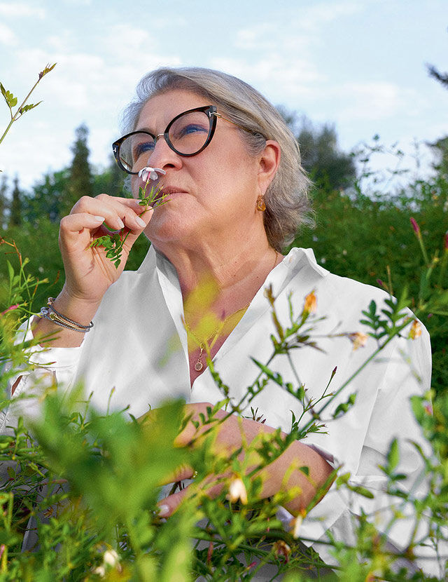 Eine Frau mit grauen Haaren und schwarzer Katzenaugenbrille steht in einem uppigen grunen Garten und riecht an einer kleinen rosa Blume, die sie sich an die Nase halt. Sie tragt ein weißes Hemd mit Kragen und Schmuck, darunter Ohrringe, eine Halskette und Armbander. Der Himmel ist strahlend blau und im Hintergrund sind einige Wolken zu sehen, was auf einen klaren Tag hindeutet. Im Vorder- und Mittelgrund sind verschiedene grune Pflanzen und einige kleine, helle Bluten zu sehen.