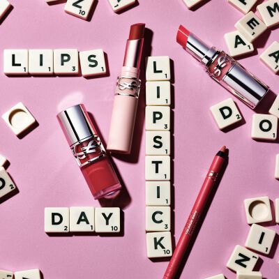 YSL Lippenstifte und Lippenkonturenstift auf rosa Hintergrund mit Scrabble-Buchstaben, die die Worter "LIPS", "LIPSTICK" und "DAY" bilden
