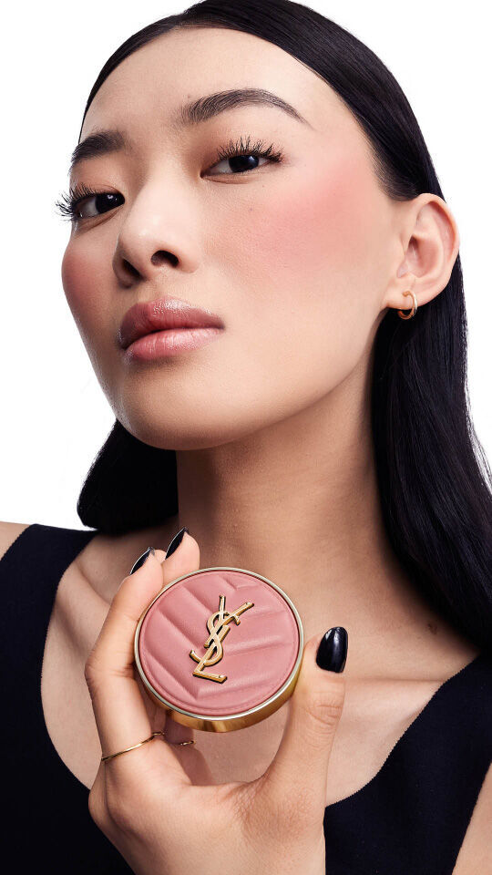 A model with long black hair holds a circular blush compact featuring the YSL logo, showcasing a soft makeup look with a hint of blush on her cheeks.