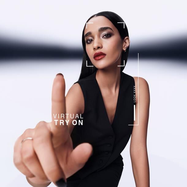 Frau zeigt nach vorne, interagiert mit virtuellem Anprobiersystem. Grafik zeigt "VIRTUAL TRY ON" und "LOOK XXXX" um ihr Gesicht. Sie tragt eine schwarze Weste.​