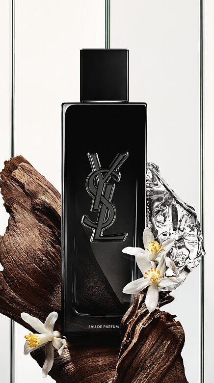 Eine schwarze Yves Saint Laurent Eau de Parfum Flasche mit dem YSL-Logo, umgeben von dunklen Holzstucken, zarten weißen Bluten und einem zerknitterten silbernen, reflektierenden Objekt.