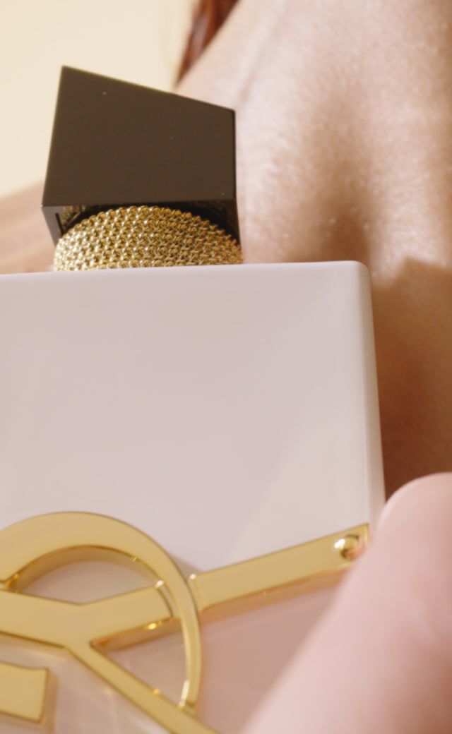 A close-up of a white perfume bottle with a gold emblem, held near a person's neck, showcasing smooth skin and a luxurious aesthetic.