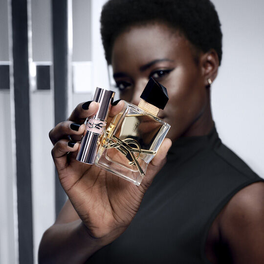 Eine Frau mit dunkler Haut und kurzen Haaren halt ein Yves Saint Laurent Parfumflaschchen und einen dazu passenden Lippenstift, beide mit dem ikonischen YSL-Logo.