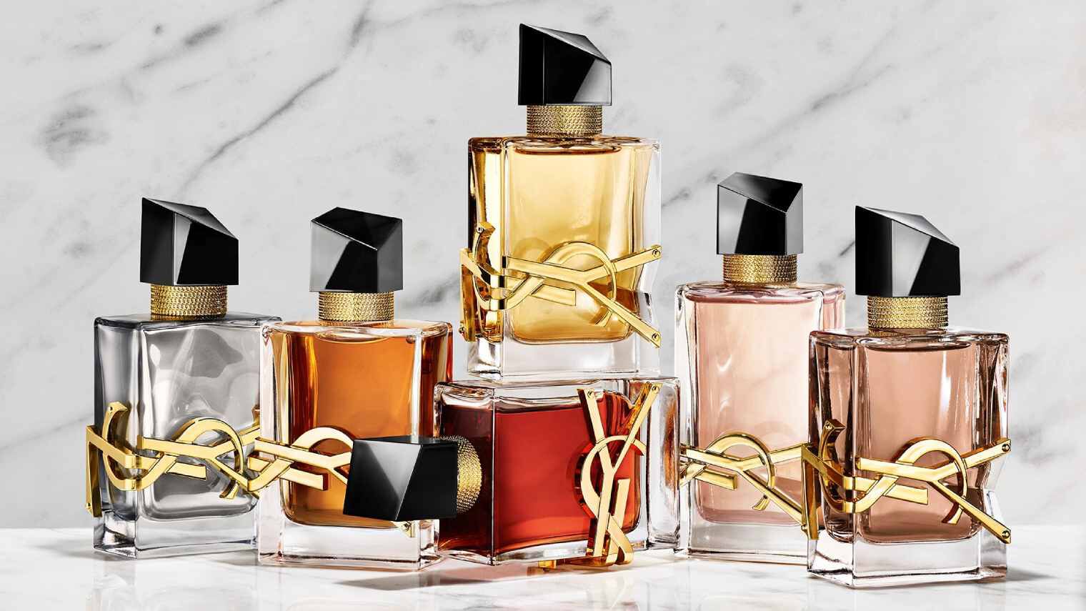 Eine Hand halt einen roten YSL-Parfumflakon neben zwei weiteren Duften auf einem Steinsockel.