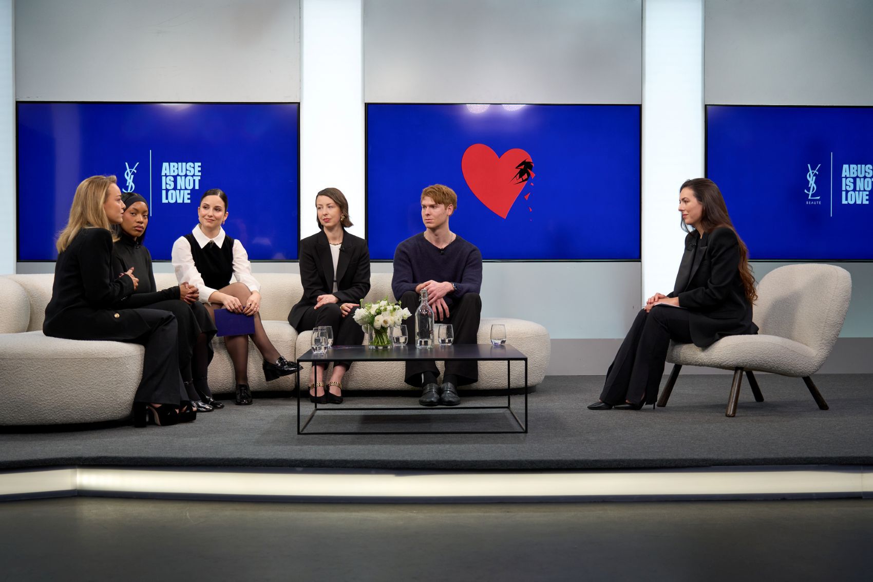Podiumsdiskussion mit sechs Sprechern (vier Frauen, zwei Manner) auf Sofas sitzend, vor großen blauen Bildschirmen mit Botschaften, in einem modernen Raum.