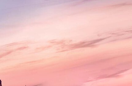 A perfume bottle with a black ribbon sits against a soft gradient sky at sunset, overlooking a cityscape.