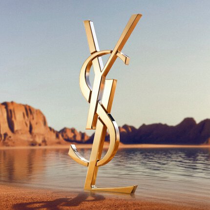 Ein großes, ikonisches 3D-YSL-Logo (Yves Saint Laurent) steht an einem Wustenstrand. Das polierte goldene Monogramm spiegelt das Sonnenlicht wider und ist vor einer ruhigen Wasserflache mit dramatischen roten Felsformationen und Wustenbergen im Hintergrund unter einem klaren Himmel positioniert.