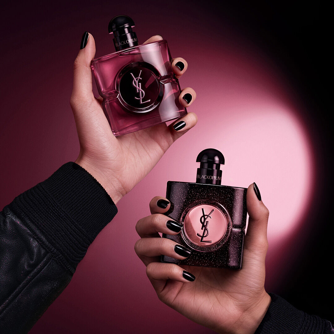 Zwei Hande halten Yves Saint Laurent Black Opium Duftflakons vor einem dunkelroten und schwarzen Hintergrund.