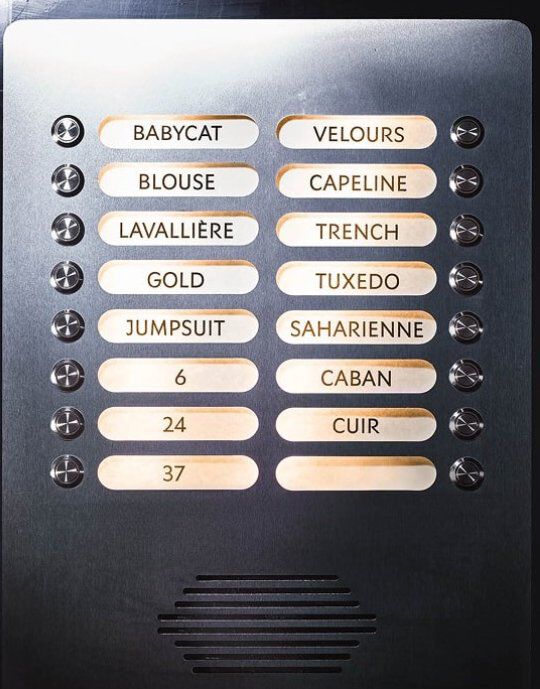 Metallplatte mit beleuchteten Knopfen, die jeweils mit einem Wort aus der YSL-Mode beschriftet sind. Zu den Beschriftungen gehoren unter anderem 'BABYCAT', 'TUXEDO', 'SAHARIENNE' und 'MUSE'. Die Platte weist kleine kreisformige Knopfe und ein Lautsprechergitter unten auf, was an ein elegantes Gegensprechanlage erinnert.