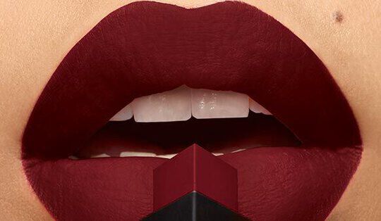 Close-up of richly colored lips with a deep red matte lipstick applied, holding a lipstick bullet in front of them.