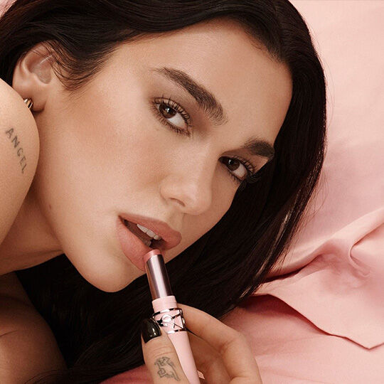 Dua Lipa tragt YSL-Lippenstift vor puderrosa Hintergrund auf.​