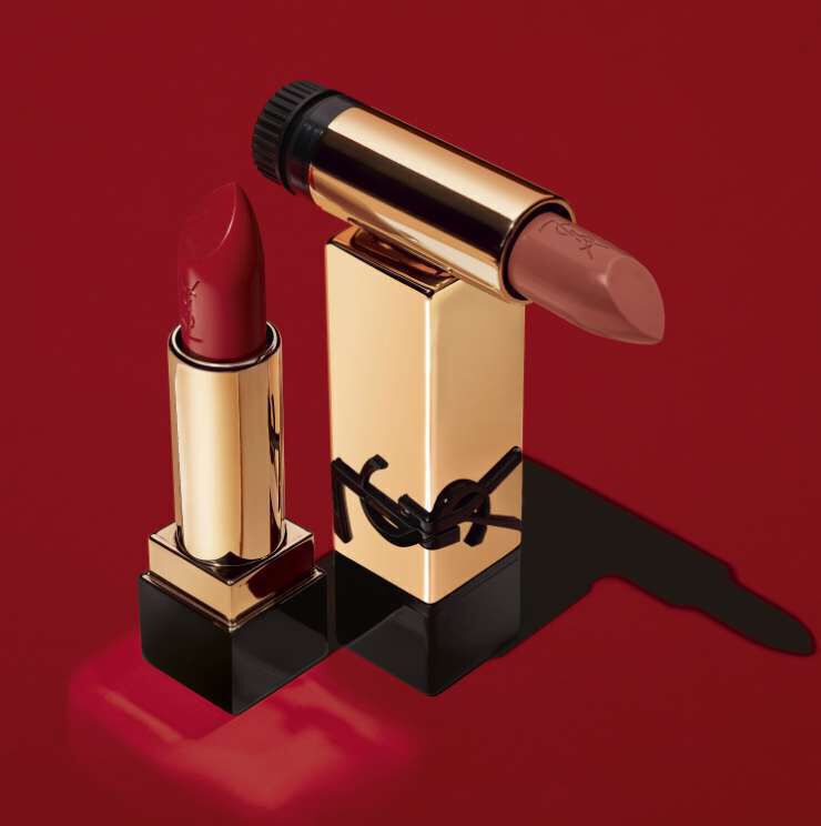 Two lipsticks are displayed against a bold red background. One is a vibrant red shade in a gold tube, while the other is a nude color surrounded by a gold casing. A portion of the nude lipstick is slightly melted, creating a glossy effect on the surface.