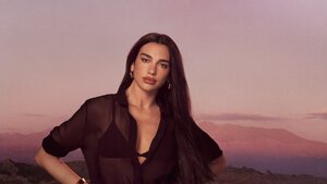 Dua Lipa posiert in einem schwarzen, transparenten Hemd vor einer bergigen Gebirgslandschaft bei Sonnenuntergang.
