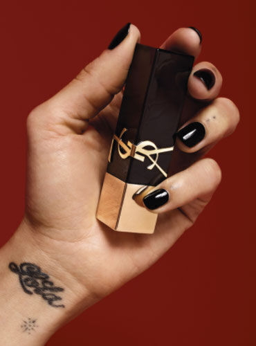A hand with black nails holds a stylish lipstick tube against a red background. The hand features a tattoo on the wrist.