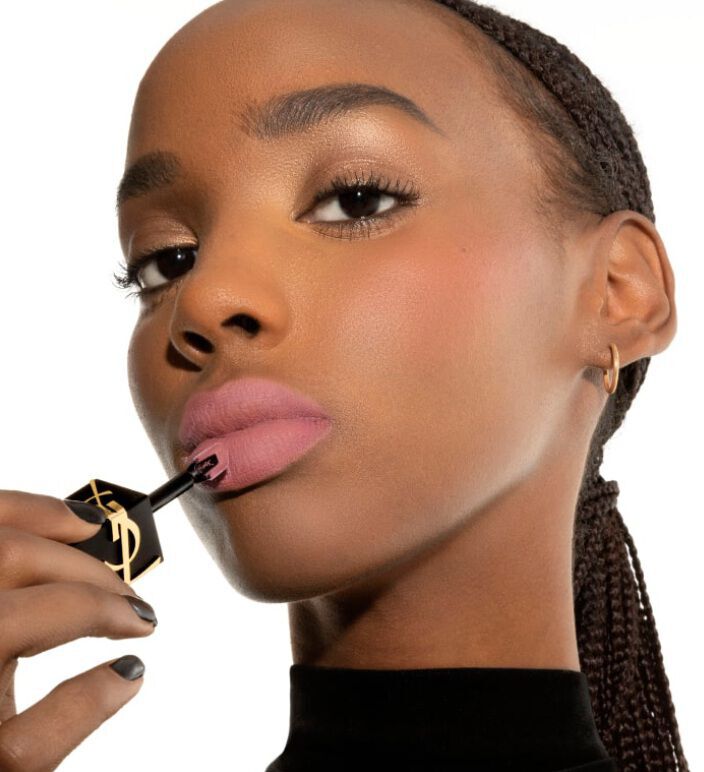 A close-up of a woman applying pink lip gloss, with her hair styled in braids, wearing a black top and subtle makeup.