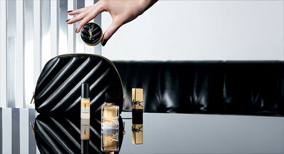 A hand is holding a compact while elegant beauty products, including a perfume and lipstick, are displayed on a reflective surface next to a stylish black cosmetic bag. The background features sleek vertical stripes.