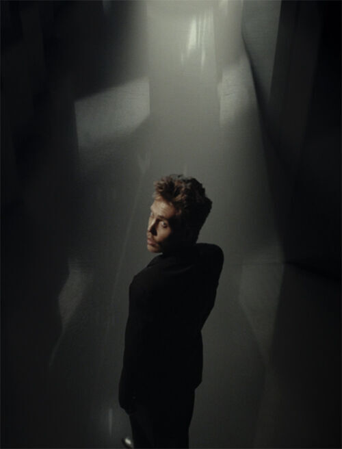 A young man in a black suit stands in a dimly lit, abstract space, looking over his shoulder with a contemplative expression. Soft light illuminates part of his face, creating a dramatic contrast with the surrounding shadows.