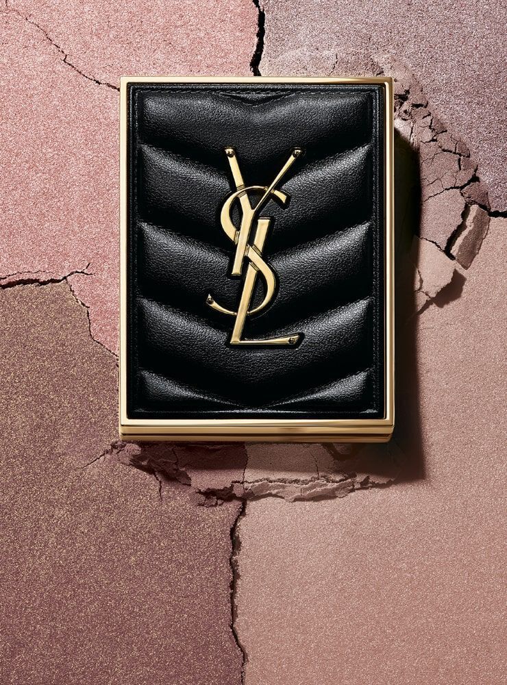 A stylish black makeup compact featuring the YSL logo, set against a background of cracked pastel colors.
