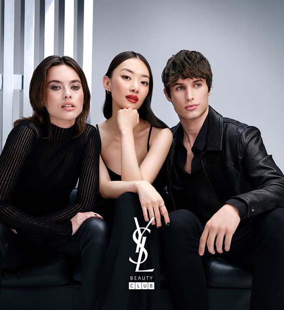 A group of three stylish models poses together in a minimalist setting. They convey a sense of confidence and fashion, with the logo "YSL BEAUTY CLUB" featured prominently at the bottom.