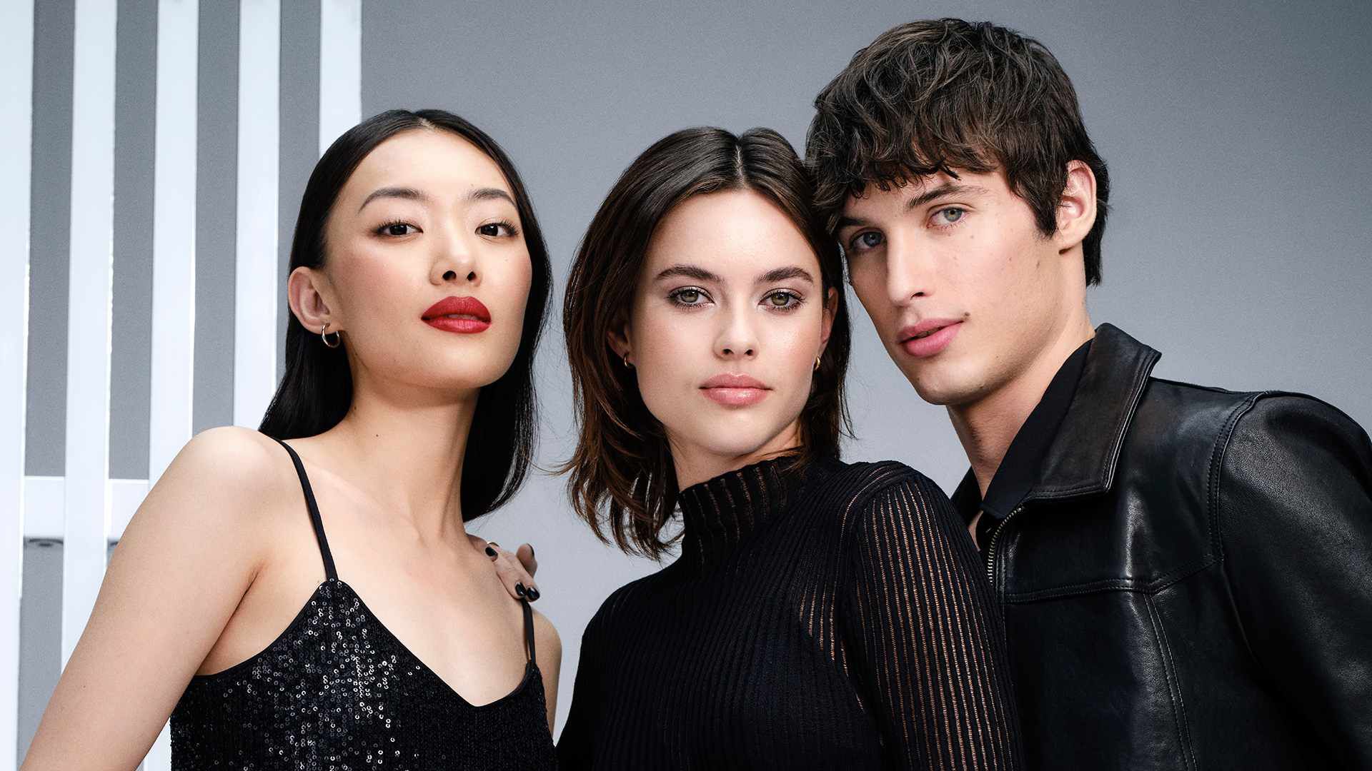 Three young adults stand close together, posing confidently. The first person on the left has long black hair and bold red lips, while the middle person features shoulder-length brown hair and a sleek black outfit. The person on the right has short, tousled hair and wears a black leather jacket, all against a neutral background.