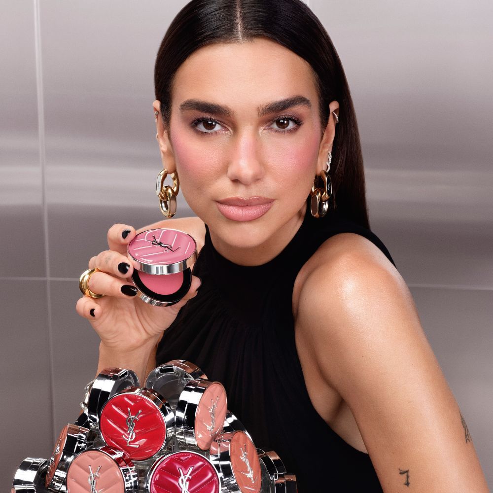 Dua Lipa halt ein geoffnetes YSL Make Me Blush Lifted Glow Cream Blush Kompakt in der Hand, wahrend die neue Kollektion der Nuancen vor ihr ausgebreitet liegt.