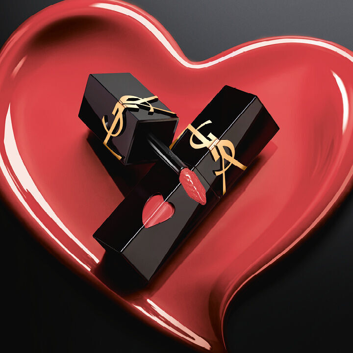 Zwei matte schwarze YSL Lip Stain Flakons mit pinkfarbenen Herz-Ausschnitten und goldenen Logos, prasentiert in einem passenden, skulpturalen matten Pink-Herz auf schwarzem Hintergrund.