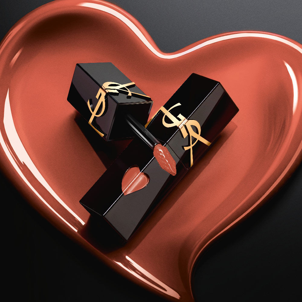 Zwei matte schwarze YSL Lip Stain Flakons mit pinkfarbenen Herz-Ausschnitten und goldenen Logos, prasentiert in einem passenden, skulpturalen matten Pink-Herz auf schwarzem Hintergrund.