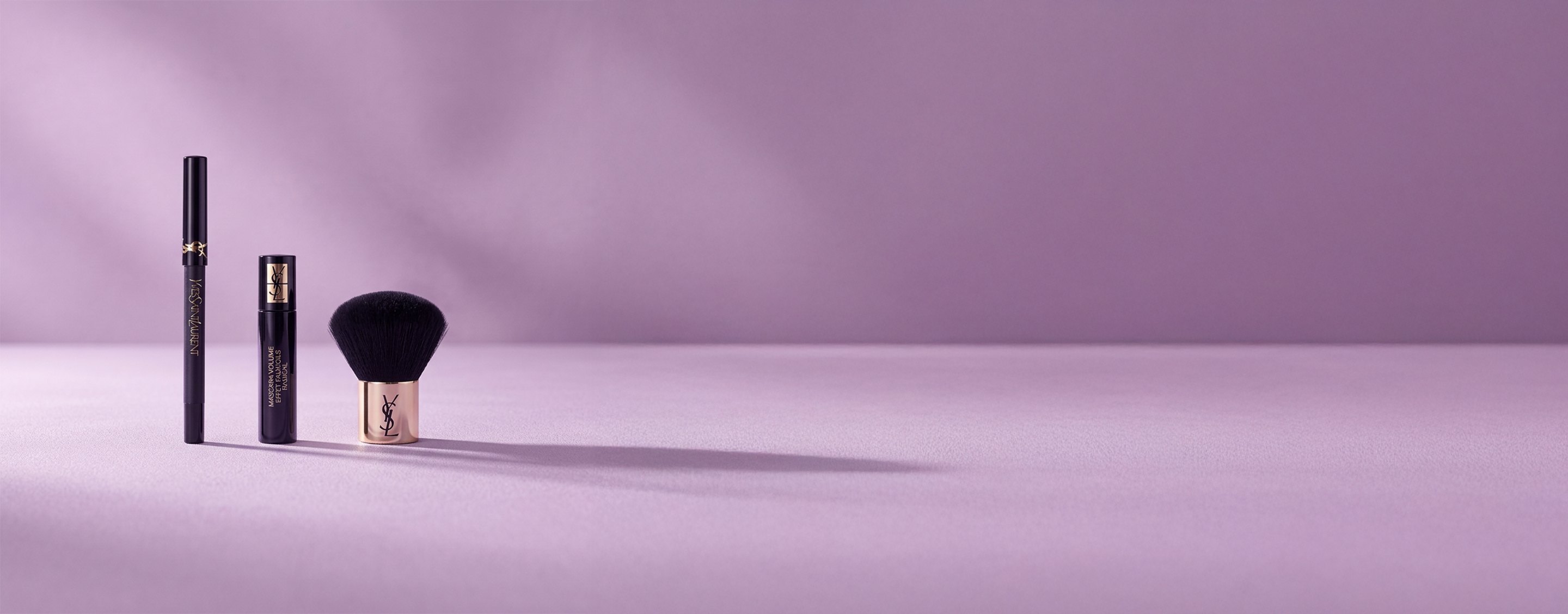 Drei Schönheitsprodukte von Yves Saint Laurent, darunter eine Wimperntusche und ein Kabuki-Pinsel, auf einem sanften lavendelfarbenen Hintergrund.