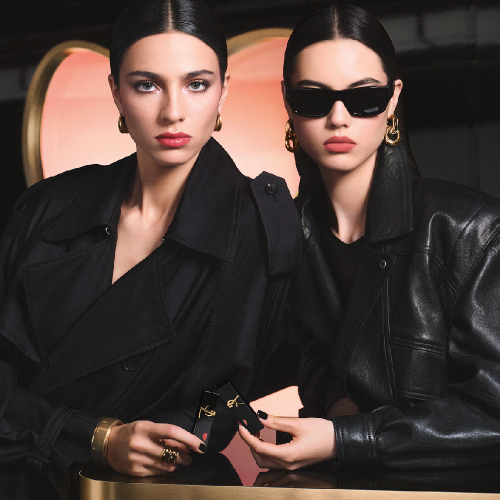 Zwei stylische Models in schwarzen Trenchcoats und Lederjacken mit YSL Lippenstiften vor einer großen pinkfarbenen Herz-Kulisse – ein glänzender Rot-Lippenlook und ein soft-matter Rosa-Lippenlook.