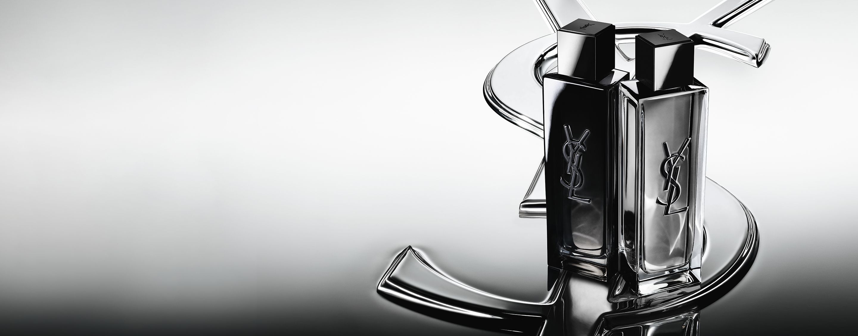 Die Flakons von MYSLF Eau de Parfum und MYSLF Eau de Toilette Intense stehen nebeneinander auf einem großen YSL-Cassandre-Logo mit spiegelndem Effekt.