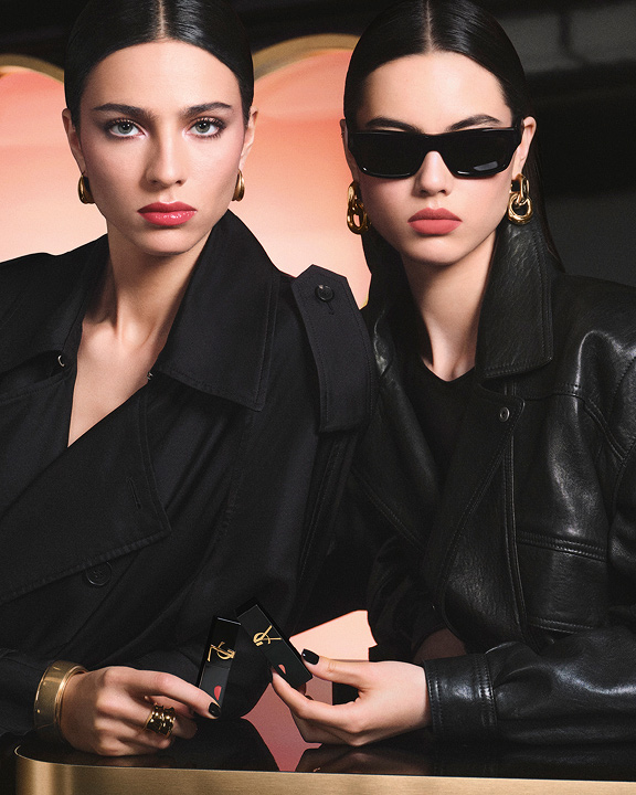 Zwei stylische Models in schwarzen Trenchcoats und Lederjacken mit YSL Lippenstiften vor einer großen pinkfarbenen Herz-Kulisse – ein glänzender Rot-Lippenlook und ein soft-matter Rosa-Lippenlook.