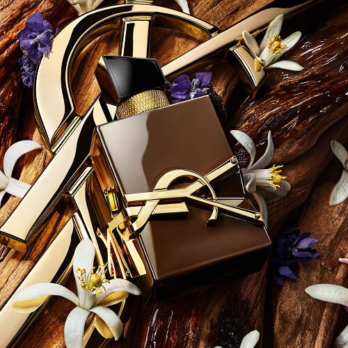 Die goldene YSL Cassandre ruht auf rohem Sandelholz, geschmückt mit weißen Orangenblüten und Lavendel, begleitet von einem Text, der die Sinnlichkeit, Schwingung und Energie des Sandelholzes heraufbeschwört.