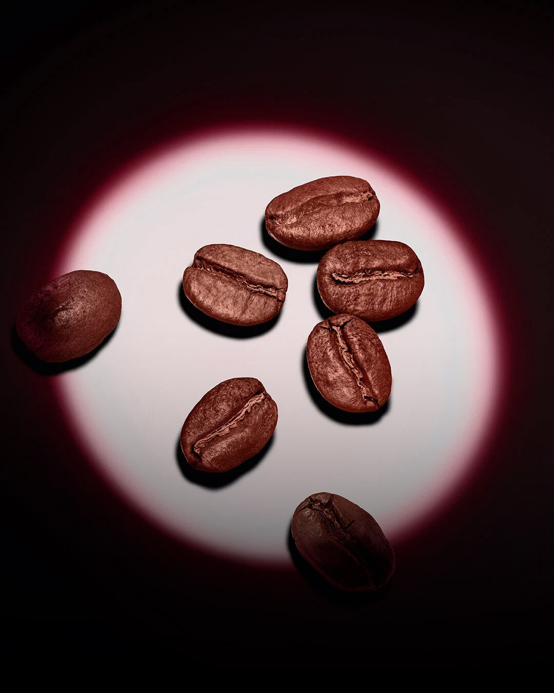 Eine Gruppe gerösteter Kaffeebohnen beleuchtet von einem pinken, kreisförmigen Spotlight auf dunklem Hintergrund. 